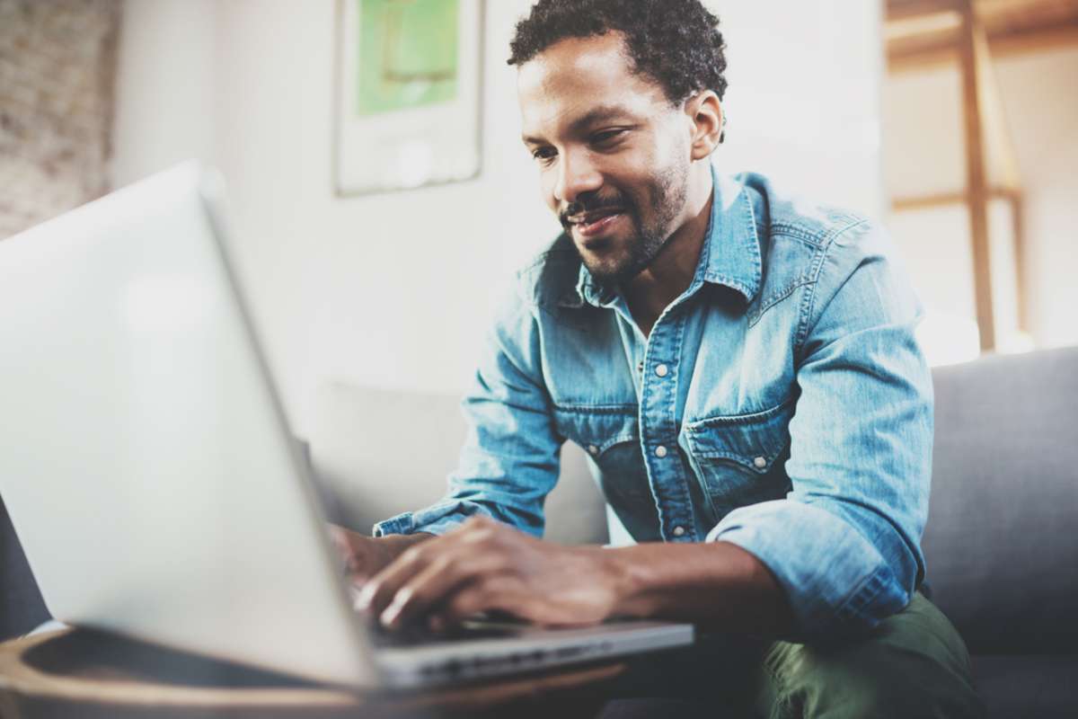 Attractive bearded African man spending free time in sofa and using laptop at modern home-1