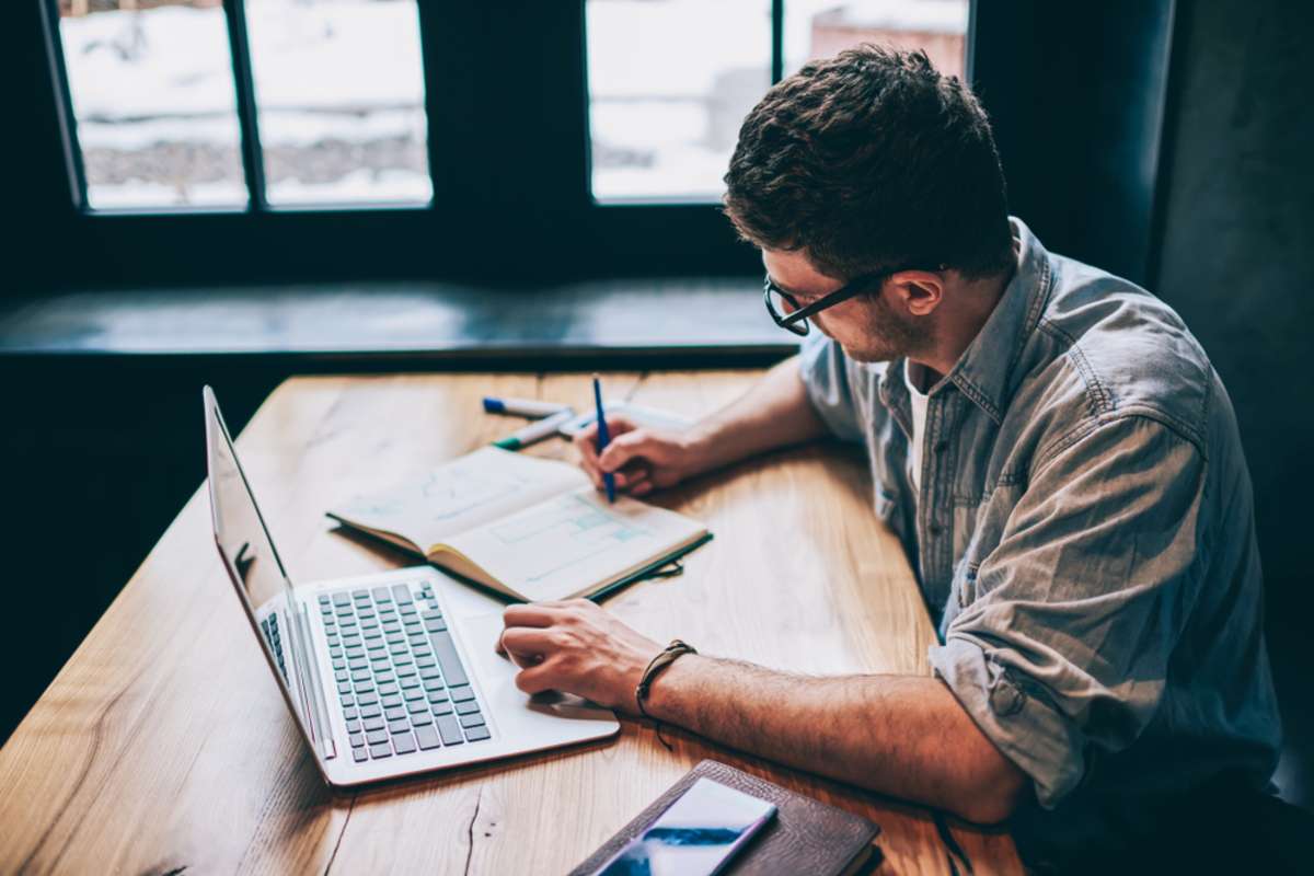 Casual dressed hipster guy writing organisation plan in textbook for education sitting at desktop with netbook