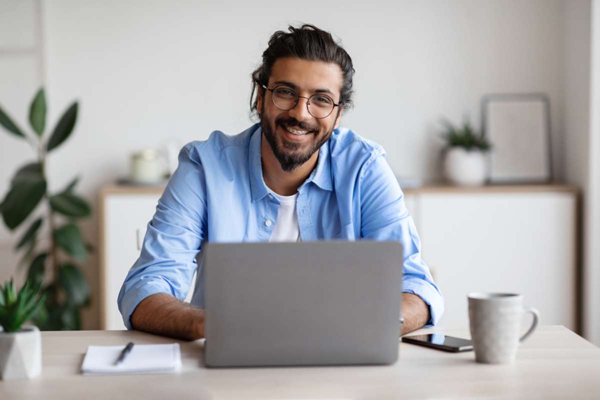 Happy Millennial Indian Man Sitting At Desk With Laptop