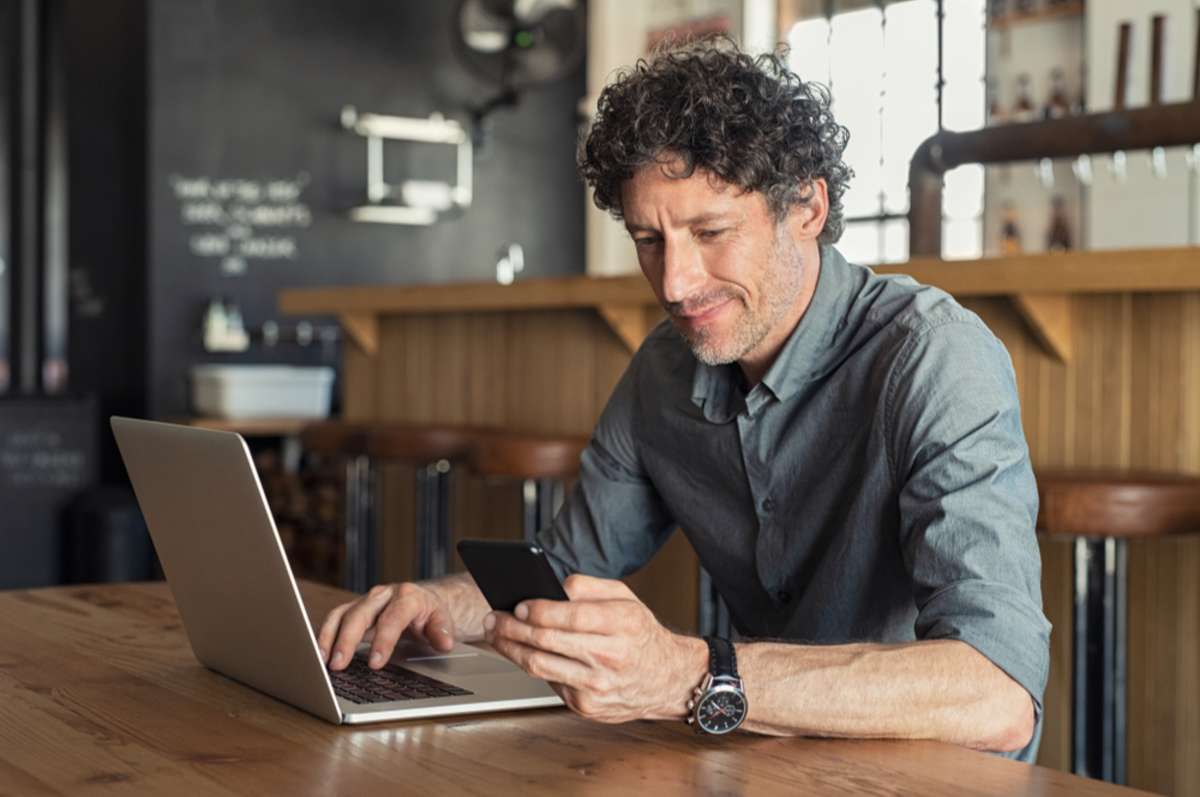 Happy mature business man sitting at cafeteria with laptop and smartphone
