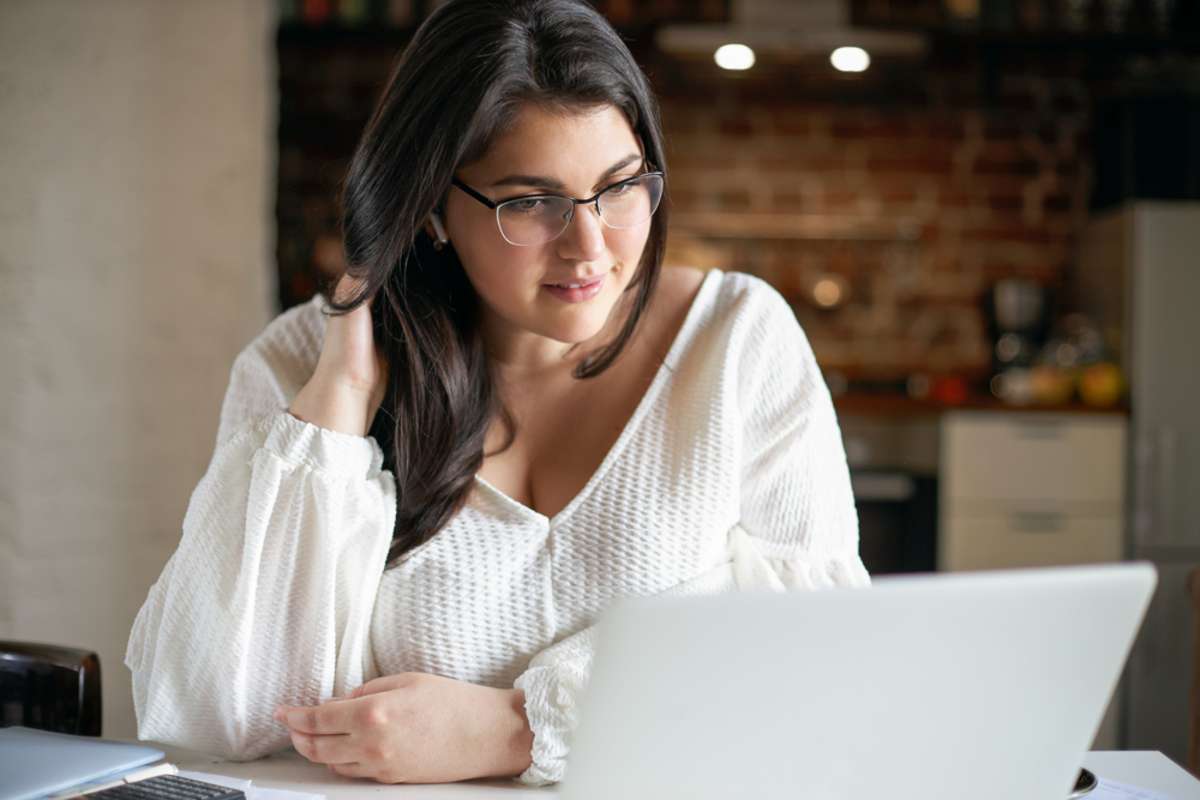 Indoor shot of attractive plus size young black haired woman in eyeglasses and white blouse sitting in front of open laptop watching video course online using wireless earplugs