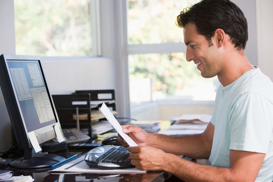 Man in home office using computer holding paperwork and smiling