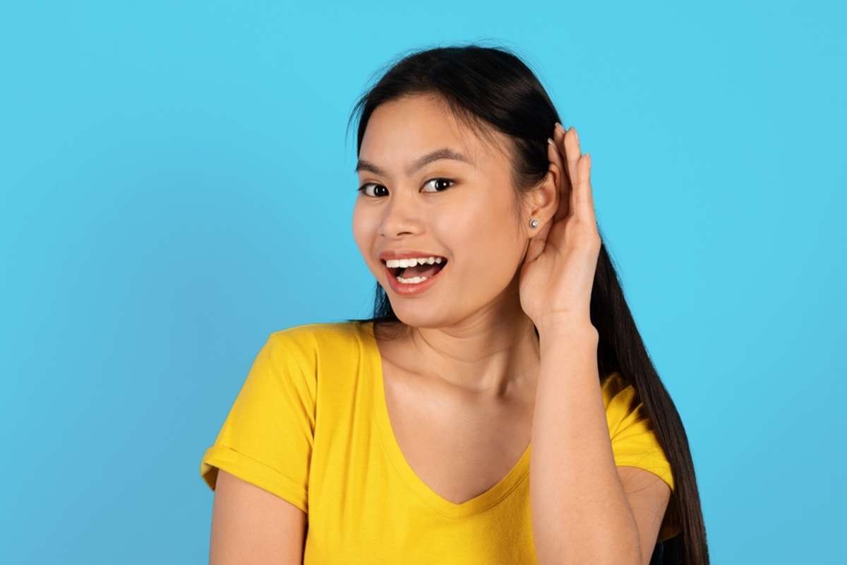 Portrait of happy inquisitive young korean lady in yellow t-shirt sticks out ear and listens news