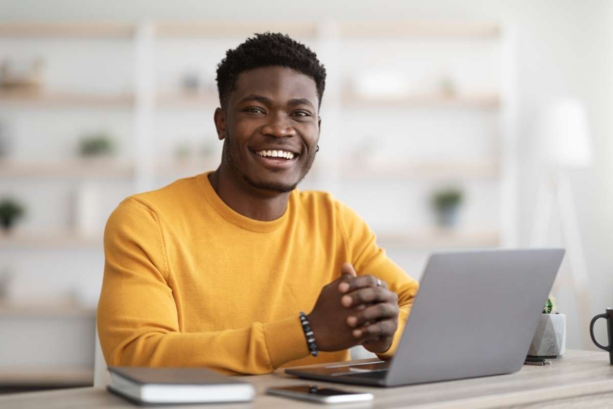 Portrait of positive young black guy in yellow sweater sitting at table