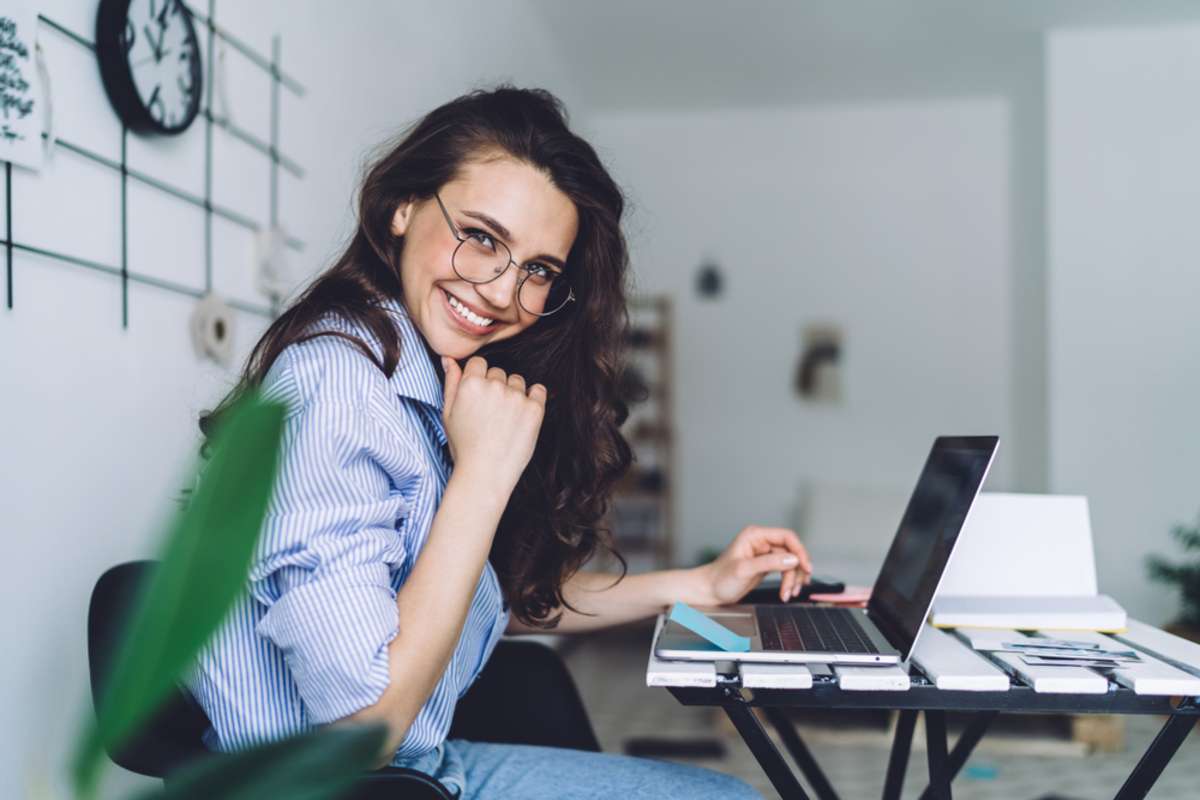 Side view of smiling long haired female in glasses in striped blue shirt sitting at laptop in workplace at home