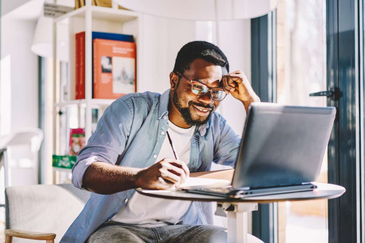 Smiling tired black guy watching on laptop and leaning with head on hand while taking notes on notebook during study in light room