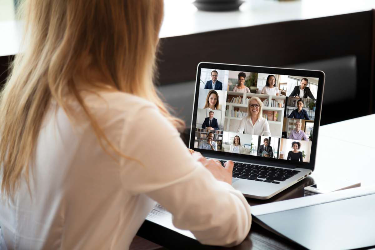 View over businesswoman shoulder sit at workplace desk looks at pc screen during group videocall with different age and ethnicity colleagues
