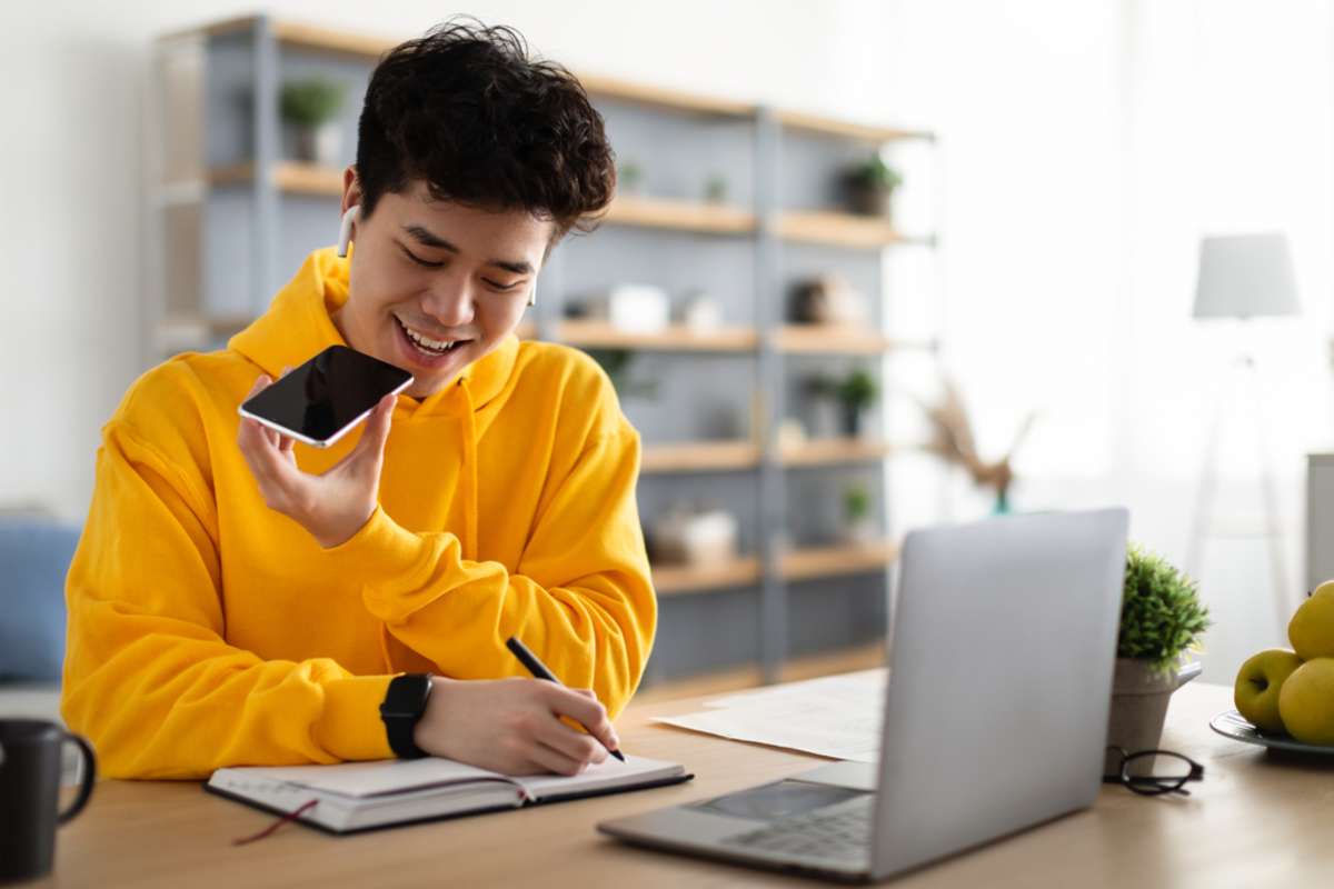 Young Asian Guy Talking On Mobile Phone And Taking Notes, Using Voice Assistant, Writing In Notebook