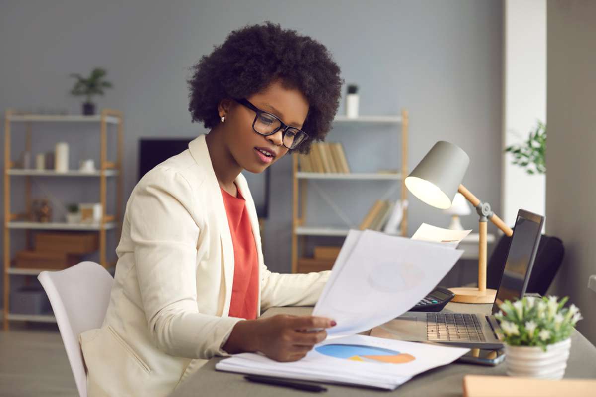 Young african american businesswoman working with laptop, financial charts and graphs reviewing data in paper documents making money work at office desk-1