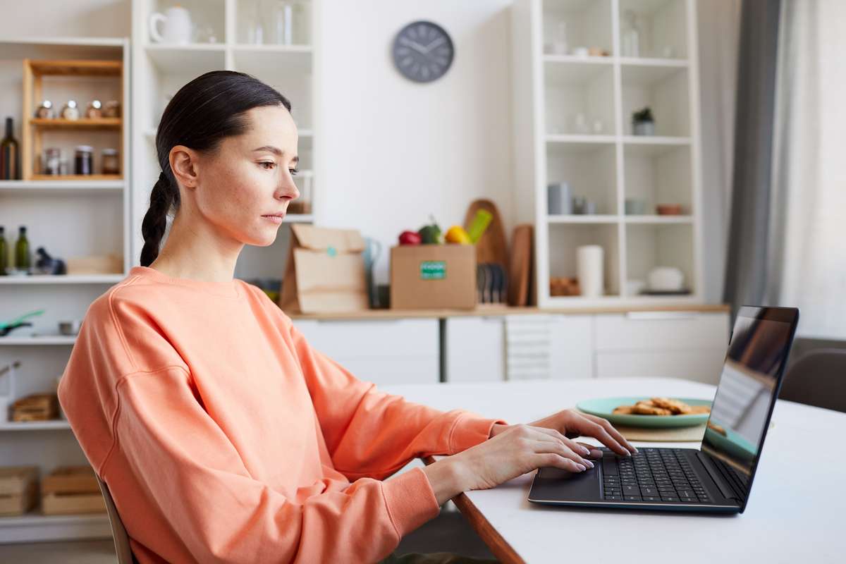 Young beautiful woman typing on laptop computer while working at the table in the kitchen