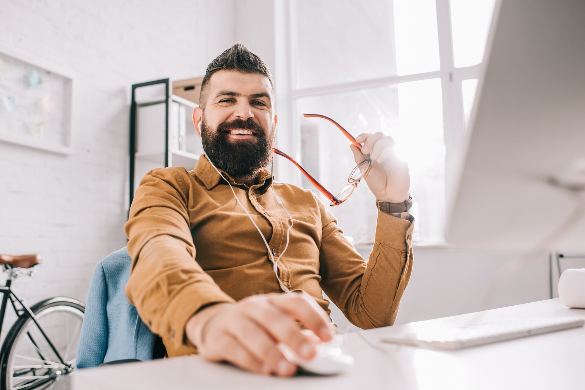 Smiling bearded adult businessman in earphones looking at camera and working at computer desk in office