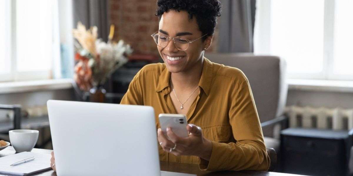 African woman sit at workplace desk holds cellphone staring at laptop